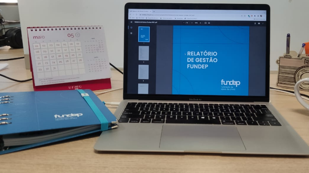 computador uma agenda azul da Fundep e do lado direito uma caneca branca com logotipo da Fundep. Ao Fundo, um calendário rosa com a logo UFMG.