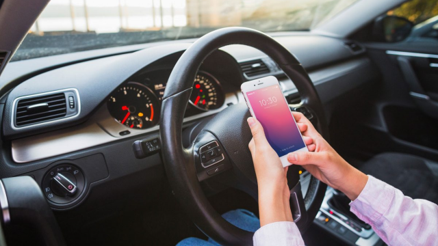 Vê-se detalhe de um painel de carro e o volante. Mãos femininas seguram um celular à frente do volante.